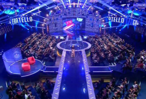 Television studio with a curved LED screen backdrop and banked audience seating under blue stage lighting