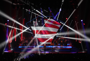 Arena concert stage with a large LED screen showing a US flag, a performer at centre stage and beams of white light over the crowd