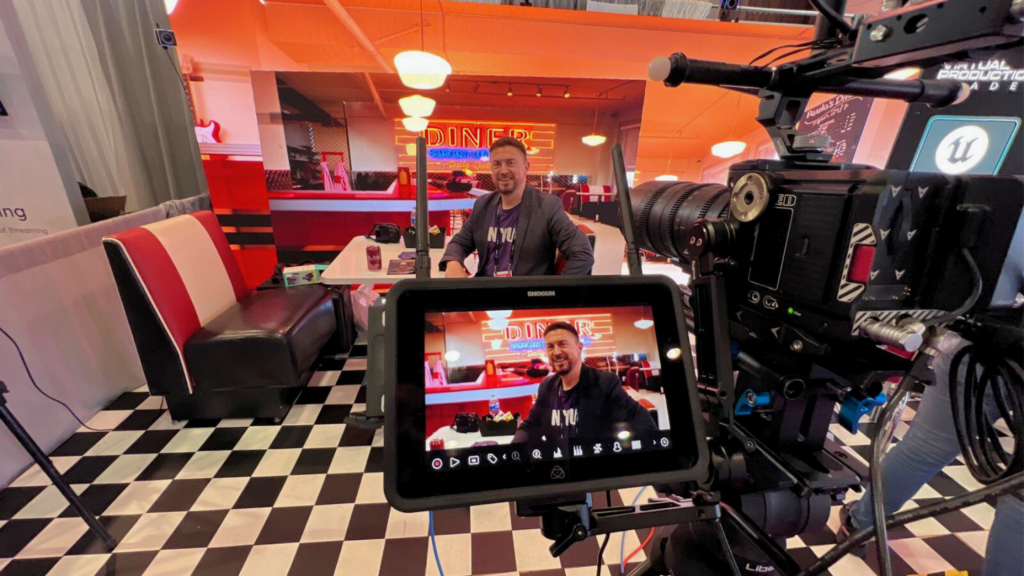 Cinema camera and monitor framing a person seated in a 1950s diner set with red booths, a checkerboard floor and a neon Diner sign