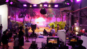Film crew and cameras on a virtual production stage in front of a large LED wall displaying a sunset beach scene