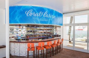 Curved LED display above the Coral Casino Beach and Cabana Club bar showing rippling pool water, with orange bar stools and an ocean view beyond