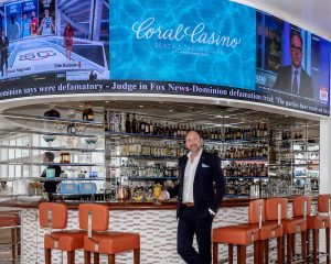 Curved LED display wrapping the Coral Casino bar in a single continuous band, running the club's branding between a live sports feed and a live news feed, with a news ticker across the full width and a guest standing at the counter